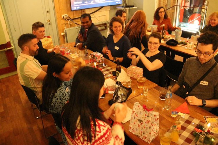 Groep van 8 mensen zit aan tafel en een van de mensen dobbelt met een dobbelsteen.